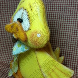 May include: A handmade yellow felt duck with orange beak, feet, and accents. The duck has a white eye, black pupil, and a small blue and white patterned bow. Gold thread tufts the top of the duck's head. The duck is a decorative item.