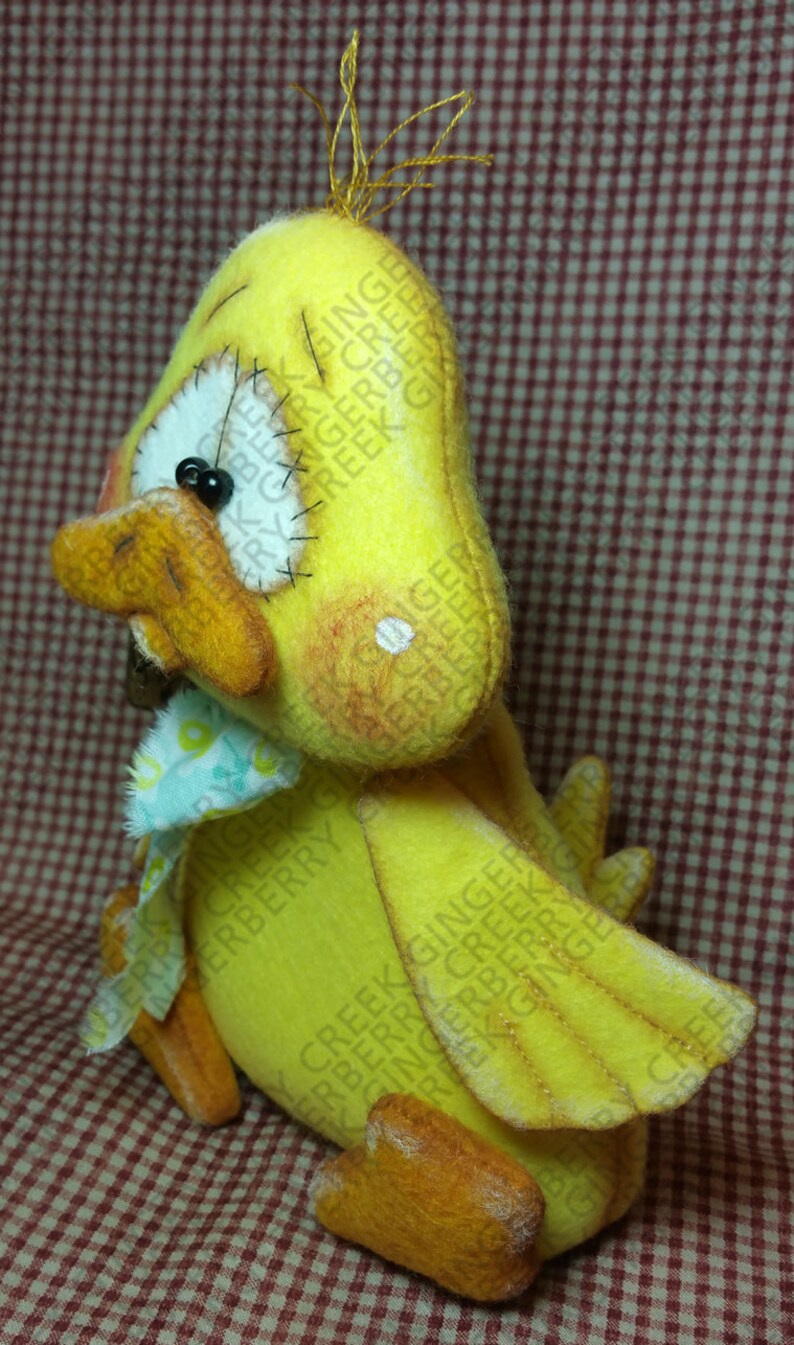 May include: A handmade yellow felt duck with orange beak, feet, and accents. The duck has a white eye, black pupil, and a small blue and white patterned bow. Gold thread tufts the top of the duck's head. The duck is a decorative item.
