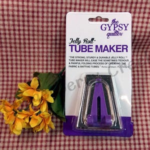 May include: A purple and silver Jelly Roll Tube Maker on a white and purple card. The card has the text "the GYPSY quilter" and "Jelly Roll TUBE MAKER". A small bouquet of yellow flowers is to the left of the card.