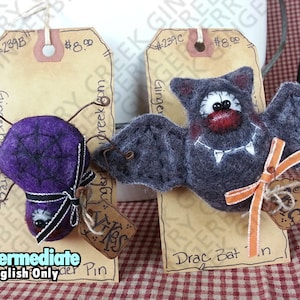 Op de afbeelding: Twee handgemaakte Halloween-viltspelden. Eén is een paarse spin met een zwarte strik, de andere is een grijze vleermuis met een oranje lint. Elke speld kost €8,00.