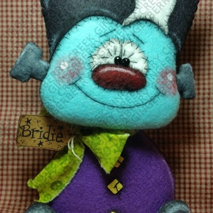May include: A handmade plush doll with a turquoise face, a black and white head, and a purple body. The doll has a stitched face with a red nose and a green scarf. A tag with the name "Bridie" is attached.