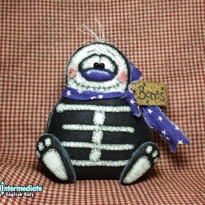 May include: A handmade Halloween skeleton decoration. The black body has white bone detailing, a purple scarf with white stars, and a tag that says "Bones". The face is white with a purple mouth and rosy cheeks.