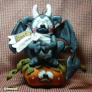 May include: A handmade Halloween decoration featuring a gray felt gargoyle with white horns and wings, perched on an orange felt pumpkin. The gargoyle has a tag that says "GDS". The pumpkin has a smiling face. The background is a red and white checkered pattern.