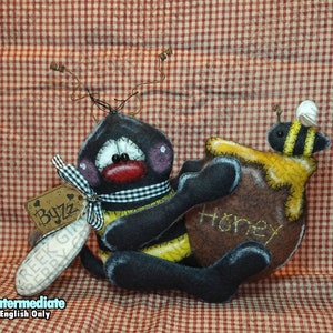 May include: A handmade felt bee doll with black, yellow, and white stripes, holding a brown honey pot with the word "HONEY" stitched on it. The bee has a black and white checkered scarf and a tag that says "Byzz".