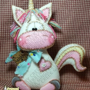Puede incluir: Un peluche de unicornio hecho a mano con cuerpo blanco, detalles rosas y azules, y un cuerno dorado. El unicornio tiene un diseño cosido, un lazo azul y una etiqueta que dice "Giddy". Sobre un fondo a cuadros rojos y blancos.