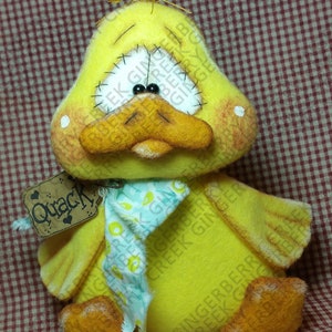 May include: A handmade yellow duck plush toy with a stitched face, black bead eyes, and an orange beak and feet. It has a small tag that says "Quack". The duck is wearing a light blue scarf with a floral pattern.