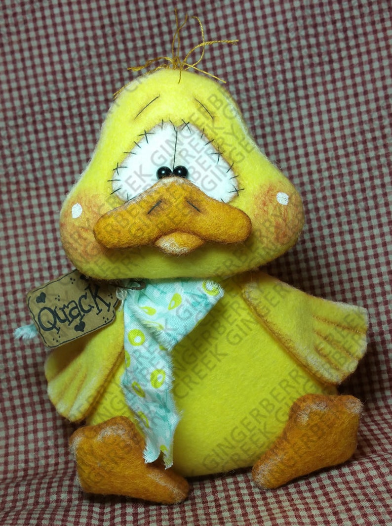 May include: A handmade yellow duck plush toy with a stitched face, black bead eyes, and an orange beak and feet. It has a small tag that says "Quack". The duck is wearing a light blue scarf with a floral pattern.