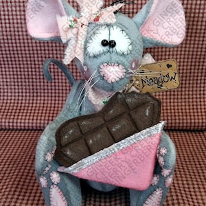 May include: A handmade gray mouse plushie with pink accents, holding a chocolate bar. The mouse has a bow, stitched details, and a tag that says "Meadow". The plushie is on a red and white checkered background.
