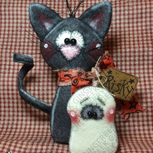 May include: A handmade Halloween cat and ghost felt ornament. The gray cat has a pink nose and a red and orange ribbon. The ghost is white with a gray face and pink cheeks. The tag reads "Risky".