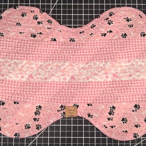 Könnte beinhalten: Ein rosa, knochenförmiges Haustierbett mit einem gesteppten Design. Das Bett weist verschiedene rosa Muster auf, darunter Vichy, Herzen und Pfotenabdrücke. Schwarze Pfotenabdrücke sind über die Ränder verstreut. Ein kleines braunes Etikett ist angebracht.