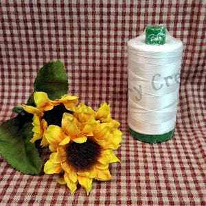Puede incluir: Un carrete de hilo blanco con bases verdes, junto a un grupo de girasoles amarillos con centros marr&oacute;n oscuro y hojas verdes. El fondo es un patr&oacute;n de cuadros rojos y blancos.