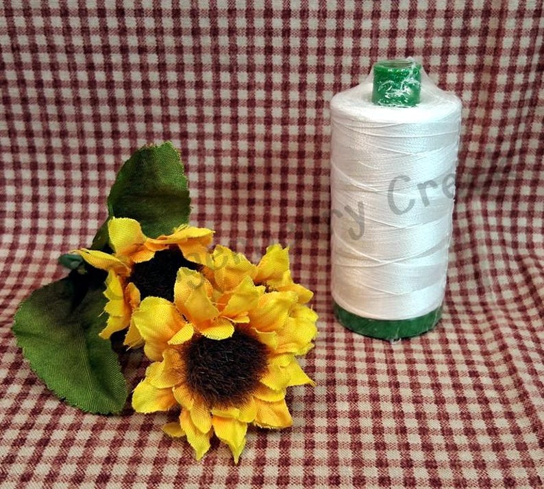 Puede incluir: Un carrete de hilo blanco con bases verdes, junto a un grupo de girasoles amarillos con centros marr&oacute;n oscuro y hojas verdes. El fondo es un patr&oacute;n de cuadros rojos y blancos.