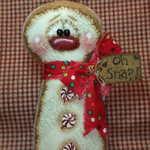 May include: A handcrafted snowman decoration with a distressed appearance. It has a red polka-dot bow, candy-style buttons, and a tag that reads "Oh Snap!". The snowman has a stitched face with black bead eyes and rosy cheeks. The background is a red and white checked pattern.