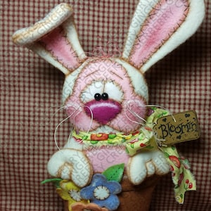 May include: A handmade Easter bunny decoration. The bunny is pink and white with stitched details, holding a pot of colorful felt flowers. A tag reads "Bloomin". The background is a red and white checkered pattern.