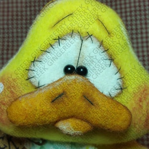 May include: A handmade, plush duck with a yellow head and orange beak. The duck has black bead eyes and stitched details, including black thread accents and white patches. The duck's face has a slightly sad expression.