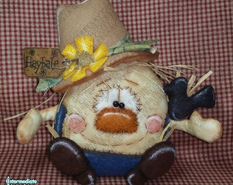 May include: A handmade scarecrow doll with a tan hat, a yellow sunflower, and a black bird. The doll has a stitched face, an orange beard, and blue overalls. A sign that says "Haybale" is attached.