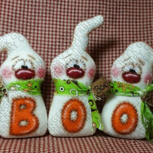 Op de afbeelding: Drie handgemaakte Halloween spookdecoraties. Elke geest is wit met een gestikt gezicht, roze wangen en een rode mond. Ze hebben oranje gestikte letters met de tekst "BOO" en groene sjaals. Elke geest heeft een label met "Eek", "Boo" en "Ahh".