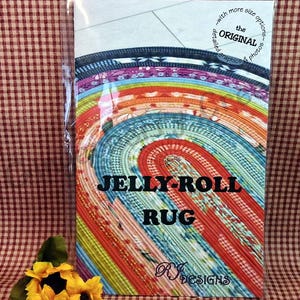 Puede incluir: Un colorido patr&oacute;n de alfombra Jelly-Roll en una funda de pl&aacute;stico. El dise&ntilde;o de la alfombra presenta un patr&oacute;n en espiral con varios colores, incluyendo rojo, azul y amarillo. El texto "JELLY-ROLL RUG" se muestra de forma destacada. Un girasol est&aacute; en la esquina inferior izquierda.