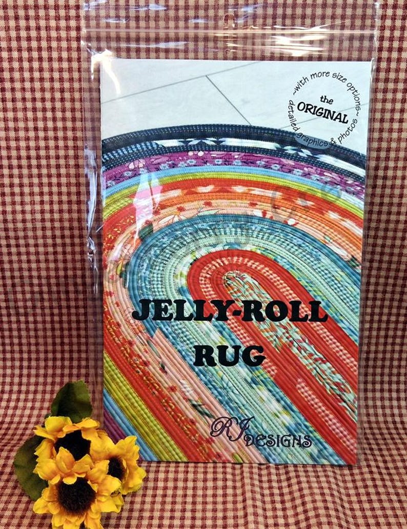 Puede incluir: Un colorido patr&oacute;n de alfombra Jelly-Roll en una funda de pl&aacute;stico. El dise&ntilde;o de la alfombra presenta un patr&oacute;n en espiral con varios colores, incluyendo rojo, azul y amarillo. El texto "JELLY-ROLL RUG" se muestra de forma destacada. Un girasol est&aacute; en la esquina inferior izquierda.