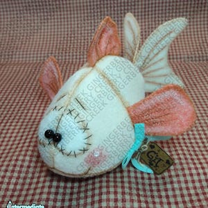 Pode incluir: Um peixe de pelúcia feito à mão com corpo creme, barbatanas pêssego e um rosto costurado. O peixe tem olhos de contas pretas e uma pequena etiqueta amarrada com uma fita azul. A etiqueta diz "Git".
