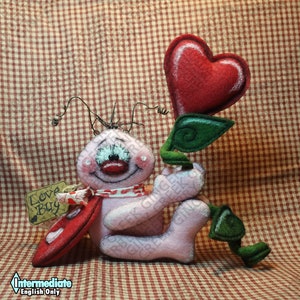 Puede incluir: Una figura rosa de fieltro de un "Love Bug" con un corazón rojo y hojas verdes. La figura tiene una cara sonriente, una bufanda roja y una etiqueta que dice "Love Bug". El fondo es un patrón de cuadros rojos y blancos.