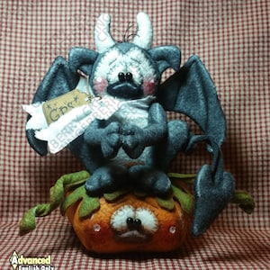 May include: A handmade felt Halloween decoration featuring a gray bat-like creature with white horns and a sad expression, perched on an orange pumpkin. The pumpkin has a stitched face and green leaves. A tag reads "GDS".