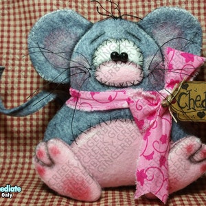 Puede incluir: Un peluche de ratón gris hecho a mano con detalles en rosa, incluyendo un lazo, nariz y pies. El ratón tiene bigotes negros y una etiqueta que dice "Cheddar". Sobre un fondo a cuadros rojos y blancos.