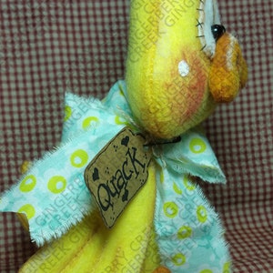May include: A handmade yellow duck figurine with a light blue and yellow patterned bow. The duck has a tag that says "Quack". The duck has orange feet and a beak. The background is a red and white checkered pattern.
