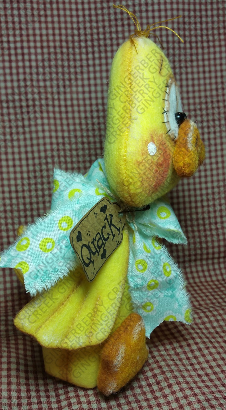 May include: A handmade yellow duck figurine with a light blue and yellow patterned bow. The duck has a tag that says "Quack". The duck has orange feet and a beak. The background is a red and white checkered pattern.