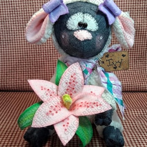 May include: A handmade stuffed sheep doll with a black face, white wool, and pink and purple accents. The doll has a pink flower and a tag that says "Ivy". The background is a red and white checkered pattern.