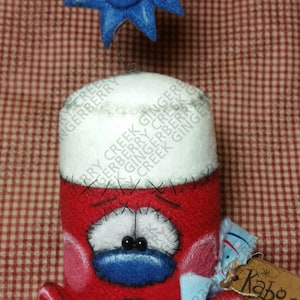 Puede incluir: Un juguete de peluche hecho a mano, rojo y blanco, con una cara y pies azules, y una estrella azul en la parte superior. El juguete tiene detalles cosidos y una etiqueta que dice "Kaboom".