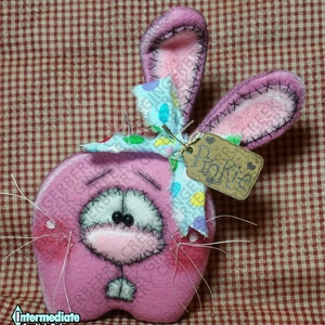 May include: A pink felt bunny with stitched details, long ears, and a whimsical expression. It has a light blue and pastel-colored bow, a tag that says "Pinkie", and white whiskers. The bunny is a handmade craft.