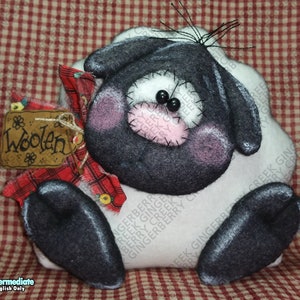 May include: A handmade sheep soft toy with a black face, ears, and legs, and a white body. It features black bead eyes, pink cheeks, and a stitched pink nose. A small wooden sign reading "Woolen" hangs from a red plaid ribbon.