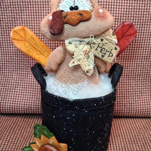 May include: A handcrafted Thanksgiving turkey decoration. The turkey is made of felt and sits in a black pot with a sunflower accent. The turkey has a tag that says "Herb". The background is a red and white checkered pattern.