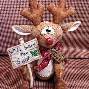 May include: A plush reindeer doll with brown fabric, large antlers, and a red nose. It wears a red and black patterned scarf and holds a rectangular sign that reads "Will Work for Santa" with a small holly accent. A tag attached reads "Redd".