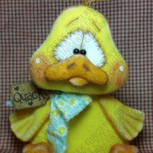 May include: A handmade yellow duck plush toy with an orange beak and feet, and black bead eyes. It has a small tag that says "Quack". The duck is wearing a blue and green scarf.