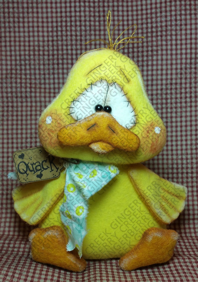 May include: A handmade yellow duck plush toy with an orange beak and feet, and black bead eyes. It has a small tag that says "Quack". The duck is wearing a blue and green scarf.