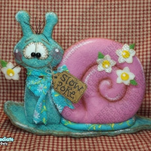 May include: A whimsical, handmade snail figurine with a turquoise body and a pink shell adorned with white flowers. The snail holds a flower and a tag that reads "Slow Poke". The design features a stitched face and a patterned scarf.