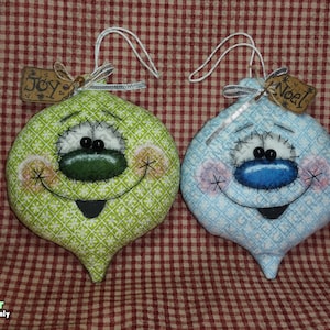 Może przedstawiać: Dwie ręcznie robione ozdoby świąteczne. Jedna jest zielona, druga niebieska, każda z uśmiechniętą twarzą i etykietą z napisem "Joy" i "Noel". Ozdoby wykonane są z tkaniny i mają wyszywany wzór.