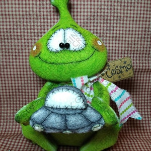 May include: A handmade green alien plush toy holding a gray spaceship. The alien has stitched features, including black button eyes and a stitched smile. A tag that says "Cosmo" is attached with a colorful ribbon. The background is a red and white checkered pattern.
