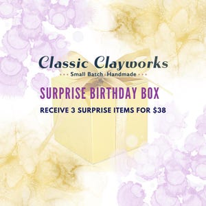 May include: A gold gift box with a cream-colored ribbon and the text "SURPRISE BIRTHDAY BOX." The box is surrounded by a watercolor-style background with purple and gold accents. The text "Classic Clayworks" is at the top.