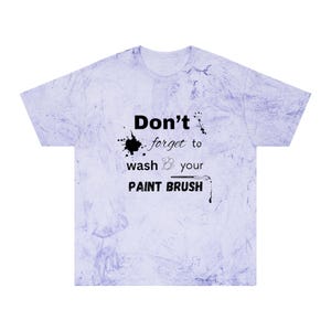 以下が含まれることがあります： 薄紫色の半袖Tシャツ。マーブル模様で、「Don't forget to wash your PAINT BRUSH」の文字が黒色でプリントされ、ペンキの飛沫のグラフィックが施されています。