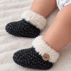 May include: A pair of black crocheted booties featuring a textured, shimmering yarn finish. Each bootie has a fluffy, cream-colored faux fur cuff and a single round, light brown wooden button attached to the side of the ankle area.