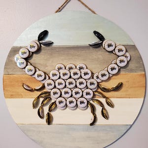 May include: A round wooden wall hanging featuring a crab design made from beer bottle caps. The crab's body is composed of numerous white and gold bottle caps, with the word "Lite" printed on them. The crab's claws and legs are made from bent bottle caps.