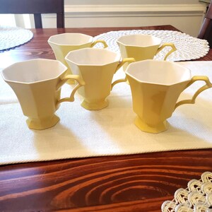 May include: Five yellow teacups with white interiors are arranged on a white runner atop a wooden table. The cups have a unique, faceted design and ornate handles. The table is made of dark wood.