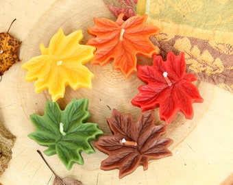 May include: Five autumn leaf-shaped candles in a circle on a wooden surface. The candles are yellow, orange, red, green, and brown.