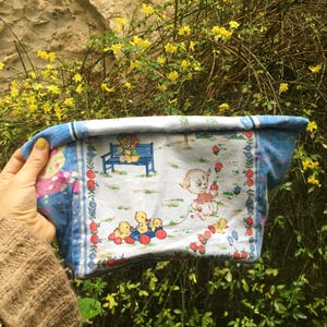 Può includere: Una borsa per bambini vintage con un motivo floreale blu e bianco. La borsa presenta una stampa affascinante di un cucciolo, anatre e un orsacchiotto su una panchina del parco. La borsa è tenuta da una persona che indossa un maglione marrone.