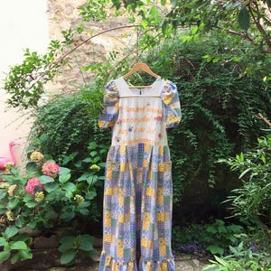 Peut inclure: Une robe longue de style vintage, ornée d'un patchwork bleu, jaune et blanc. La robe présente des manches courtes bouffantes et un corsage blanc avec des broderies et des motifs floraux. Elle est suspendue à un cintre en bois.