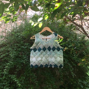 Peut inclure: Un haut sans manches avec un motif matelassé dans les tons de vert et de blanc, suspendu à un cintre en bois. Le haut présente un motif de losanges et une petite ouverture en trou de serrure dans le dos. Le fond est un buisson vert luxuriant.