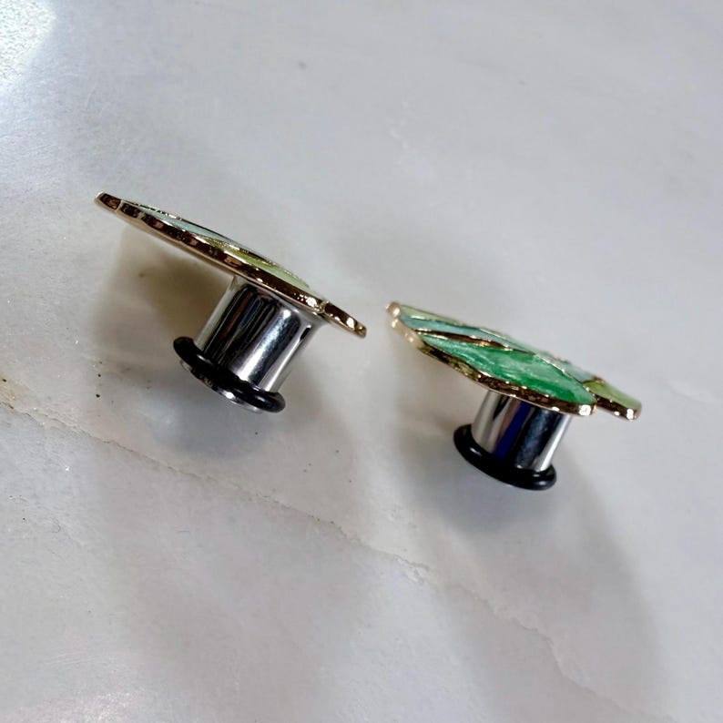May include: Two silver-colored ear plugs with a leaf design. The leaf has green and gold accents. The plugs have a black rubber ring around the base.